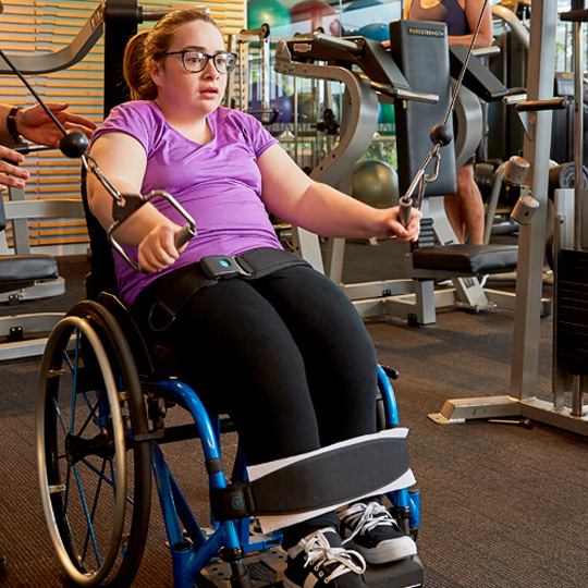 Photo of a person in a wheelchair wearing a purple shirt in a gym using a hand pull strength machine